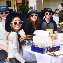 A group of smiling young adults sits at a table, each wearing white sunglasses and matching blue hats. 他们被礼品盒、小册子和插花包围着. The setting appears to be a casual event or gathering.